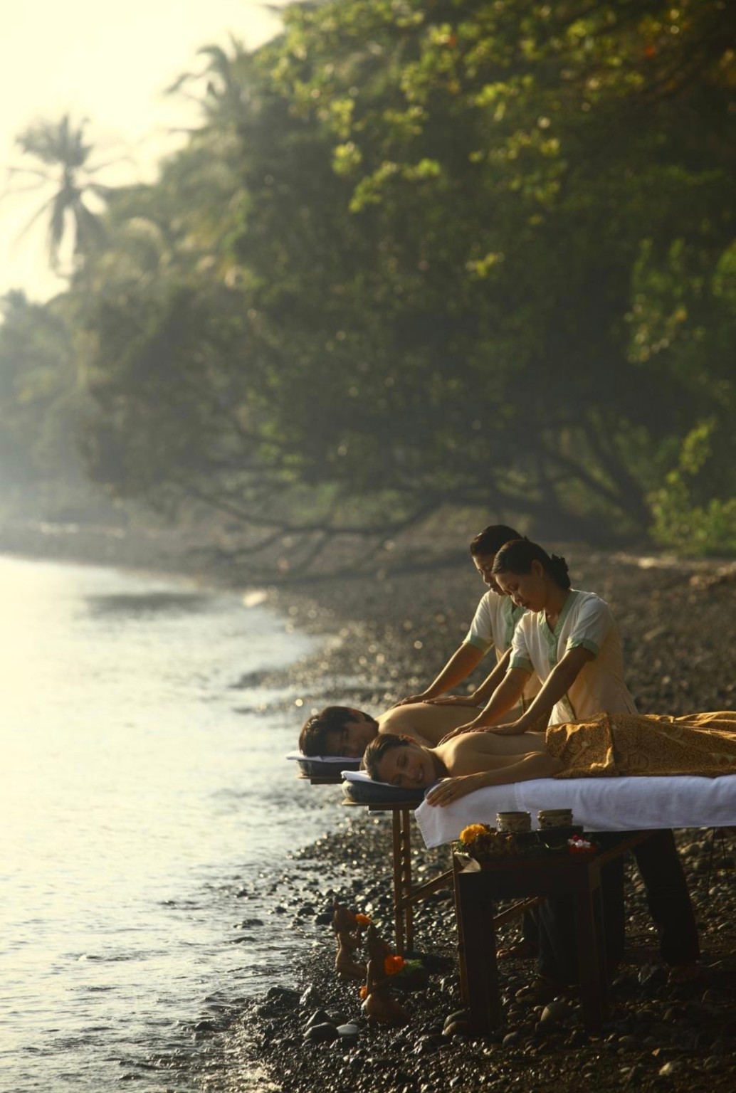 Two massage therapists treating clients beside a tropical shoreline in warm sunlight